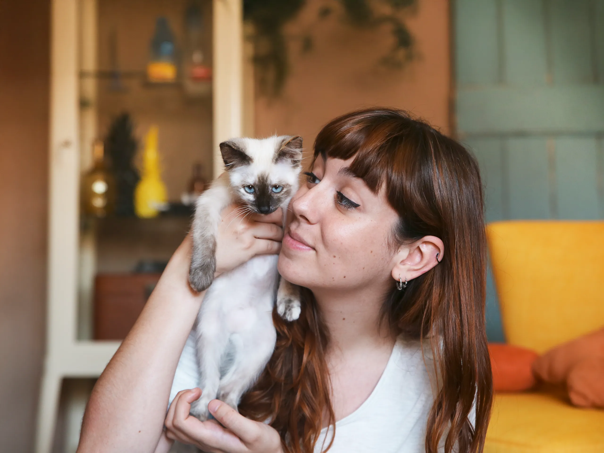 Junge Frau mit braunen Haaren hebt eine Siamkatze hoch und lächelt.