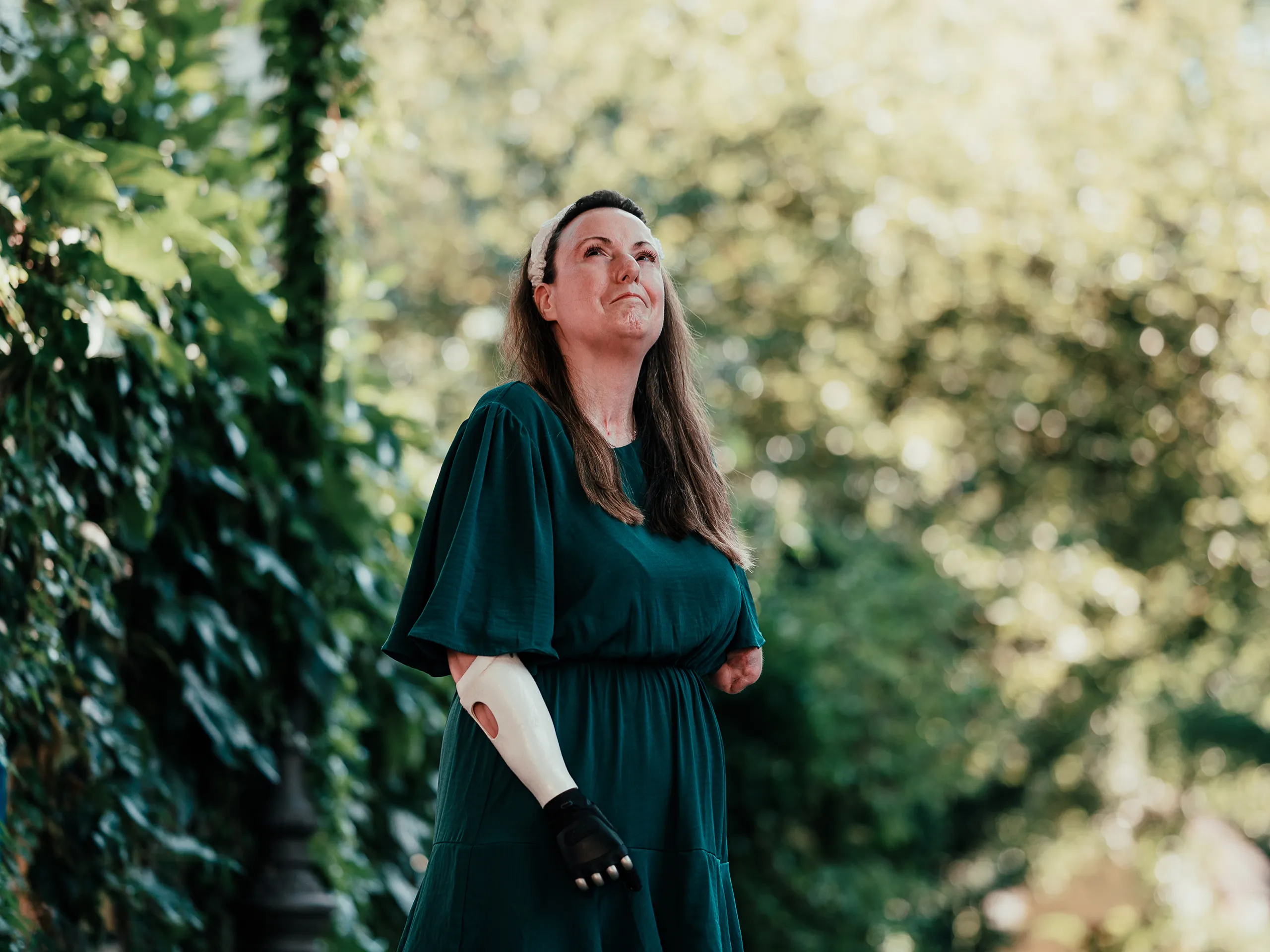 Frau mit Prothese steht im Freien, umgeben von grünen Blättern, und blickt hoffnungsvoll nach oben.