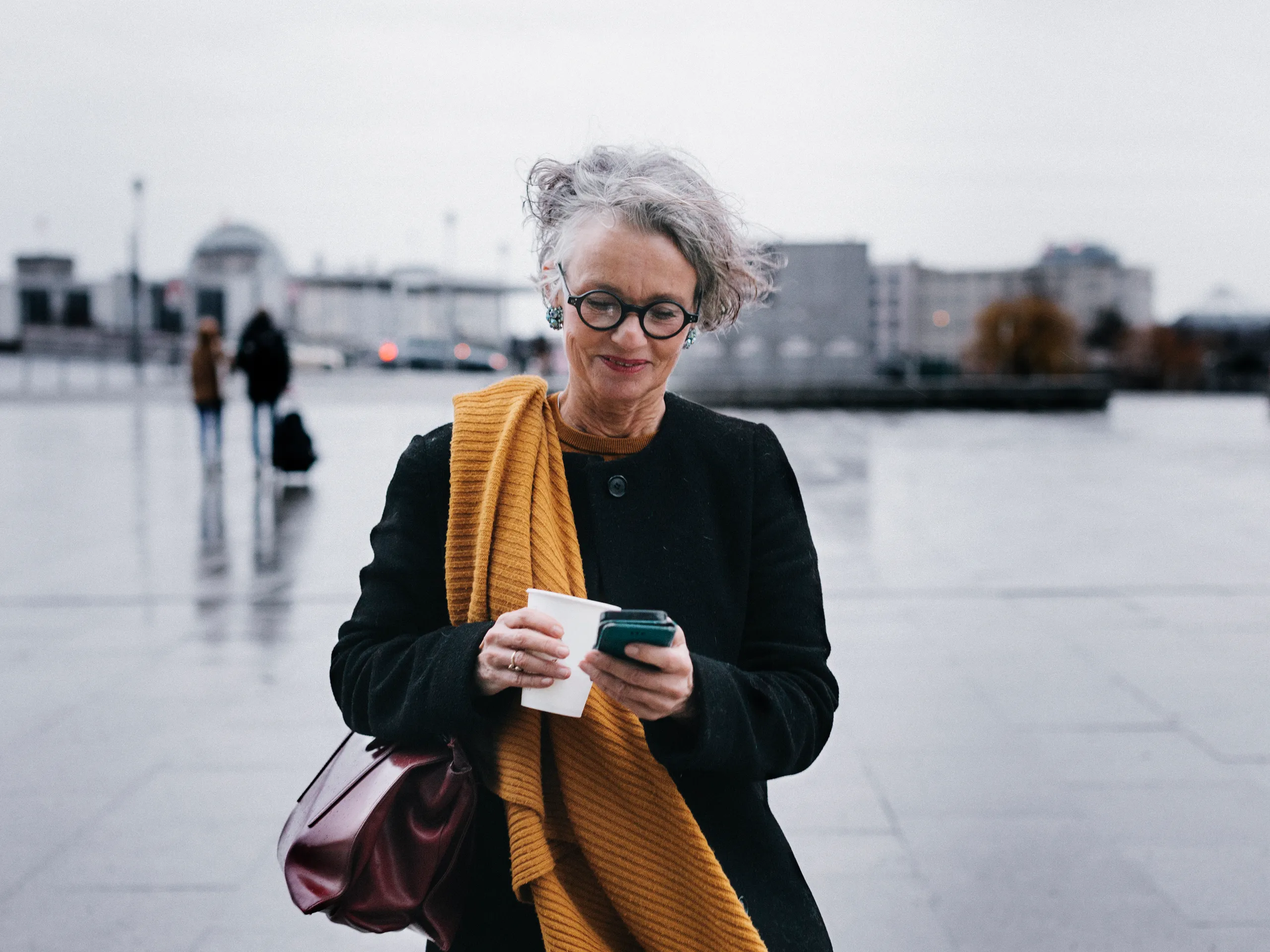 Ältere Frau mit Brille und gelbem Schal steht auf einem Platz und schaut auf ihr Smartphone