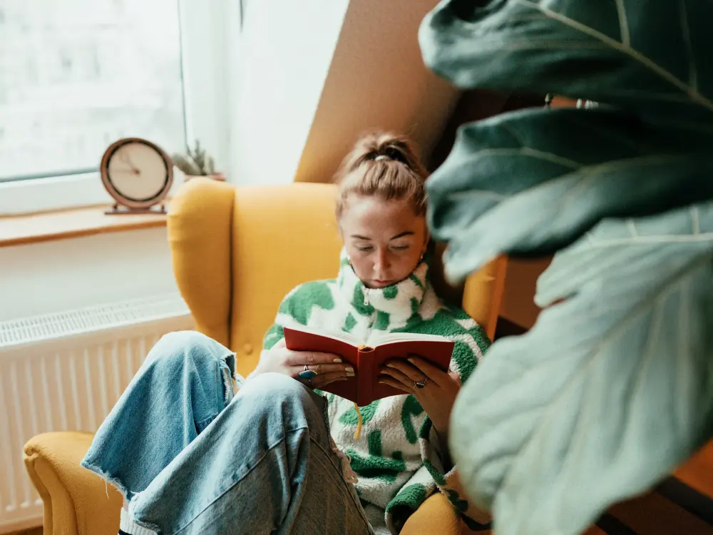 Frau sitzt in einem gelben Sessel und liest ein Buch in gemütlicher Atmosphäre