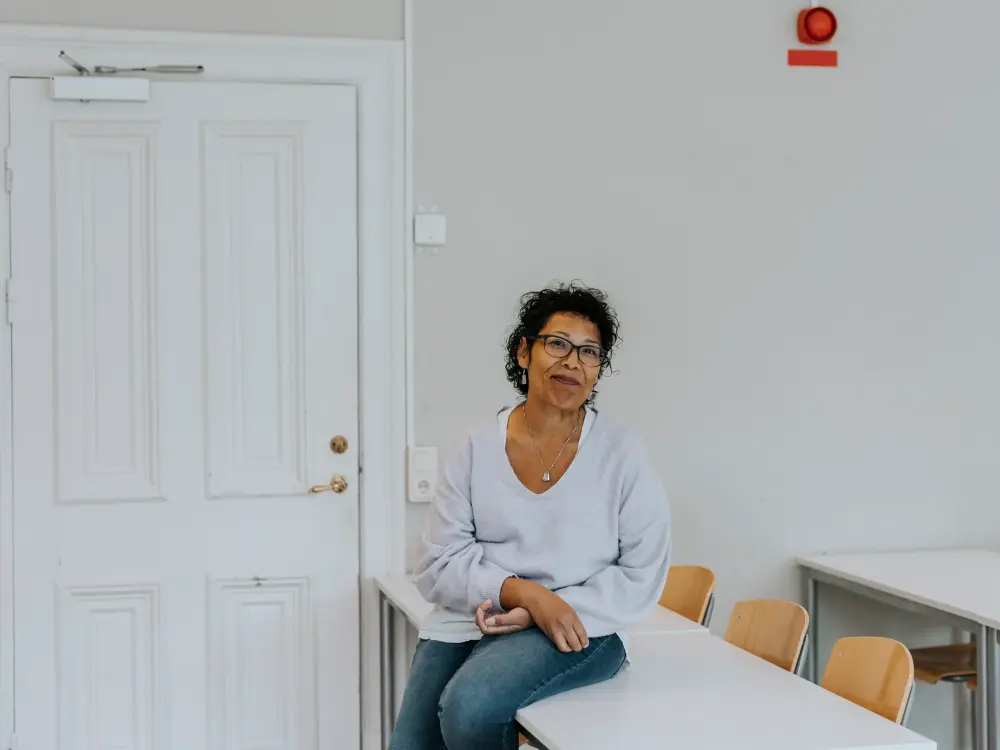 Frau mit Brille sitzt entspannt auf einem Tisch in einem leeren Klassenzimmer