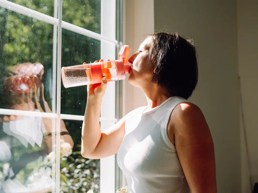 Frau trinkt aus einer Wasserflasche vor einem Fenster