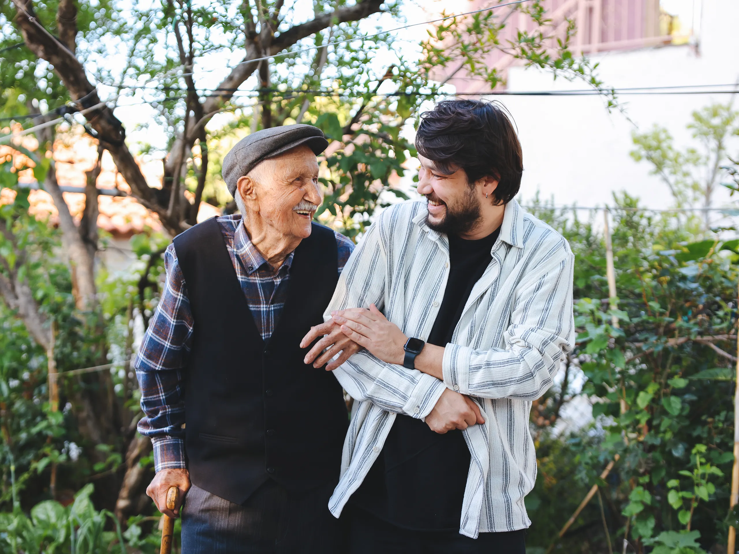Junger Mann mit Bart und älterer Mann mit Schiebermütze lachen gemeinsam im Garten.