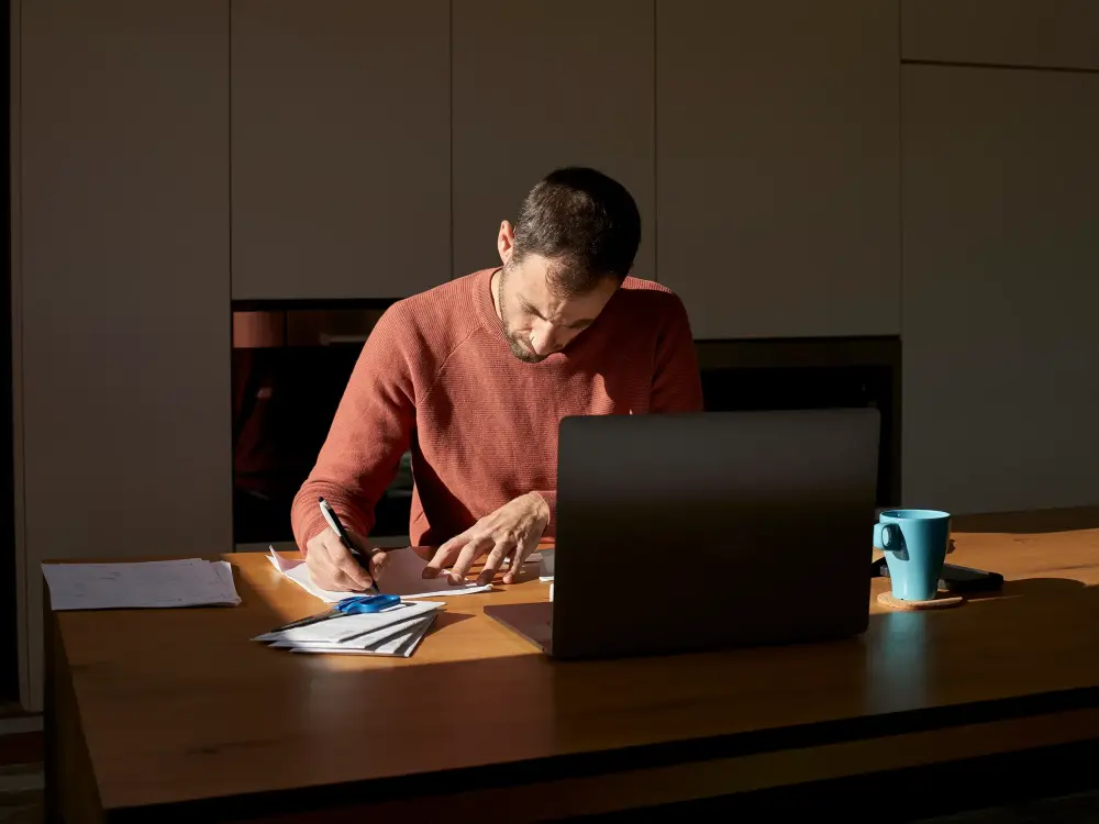 Mann arbeitet konzentriert mit Laptop und Unterlagen an einem Holztisch