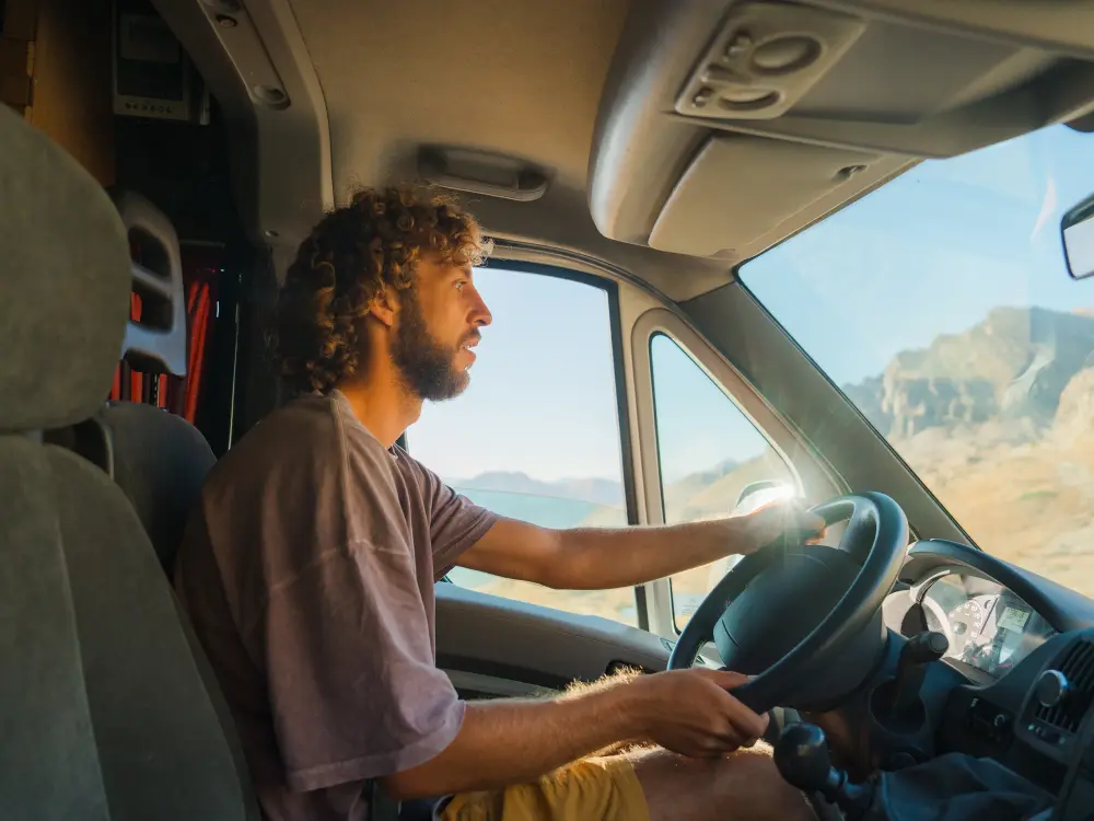 Mann mit lockigen Haaren fährt einen Camper durch eine sonnige Berglandschaft.
