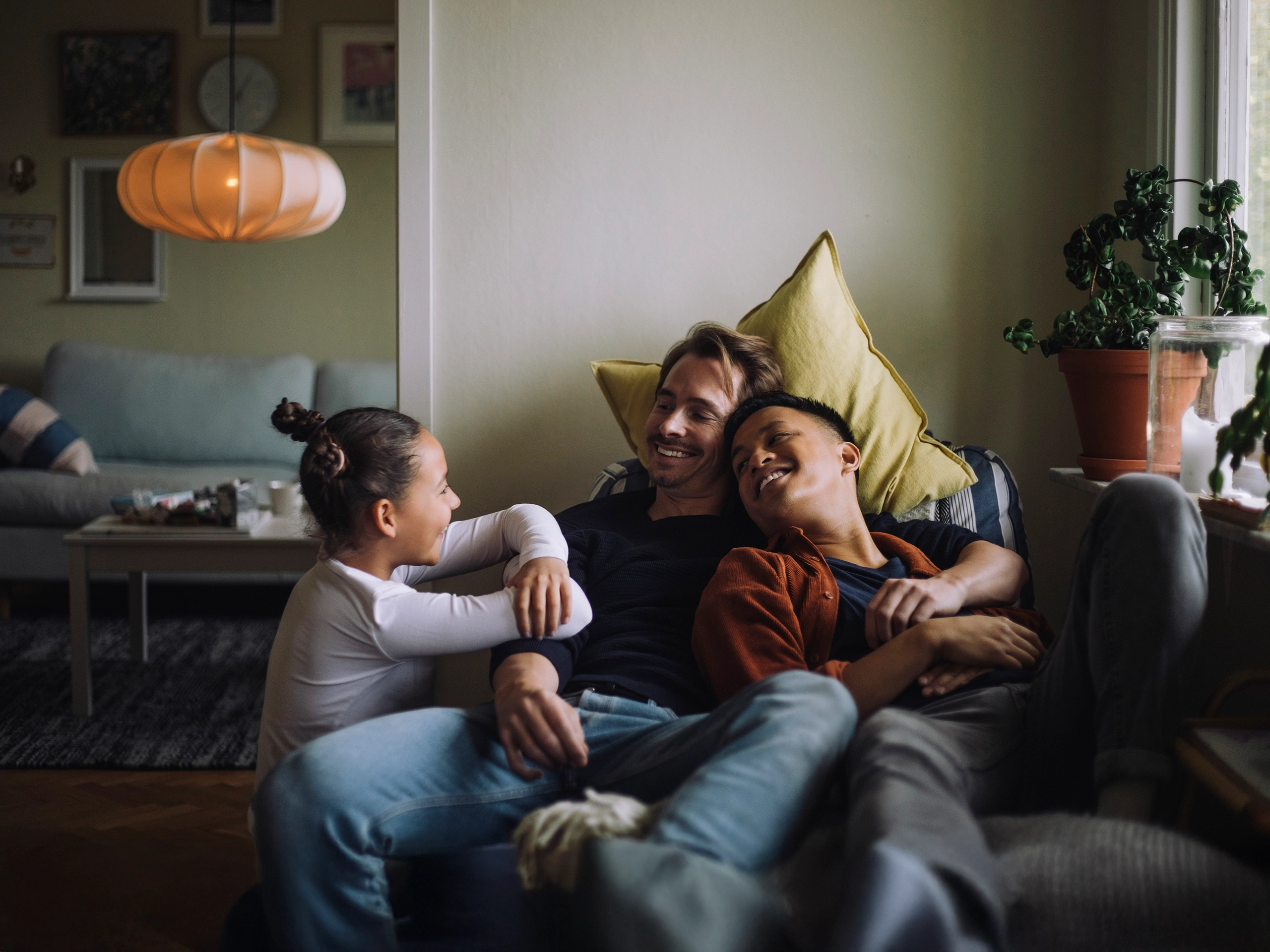 Zwei Väter kuscheln mit Tochter lachend auf dem Sofa